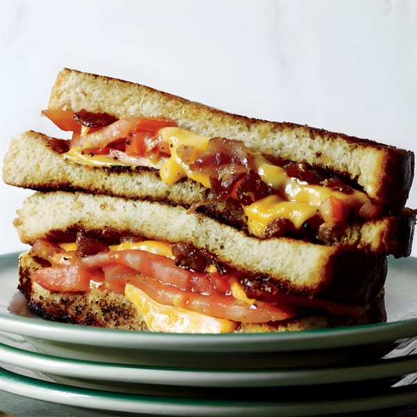 A stacked grilled cheese sandwich with melted cheese, bacon, and tomatoes on toasted bread.