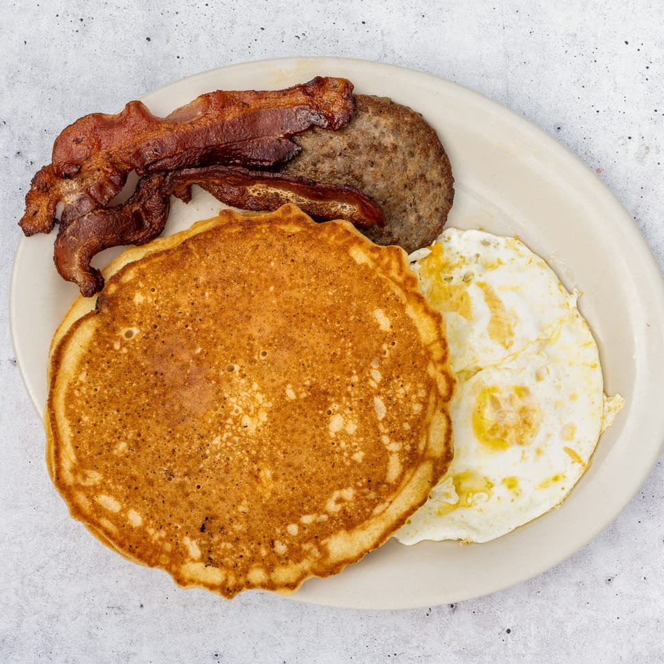 A breakfast plate with pancakes, crispy bacon, sausage patty, and two fried eggs.