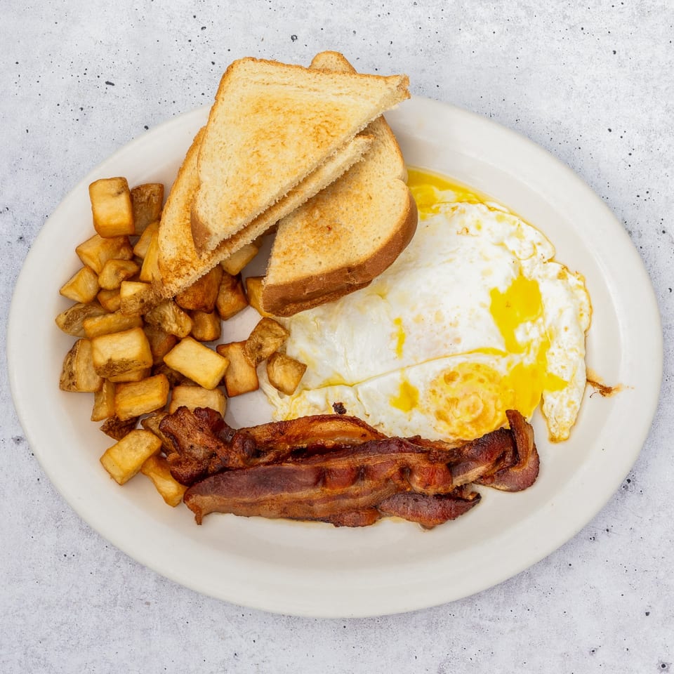 A breakfast plate with crispy bacon, fried eggs, golden home fries, and toasted bread.