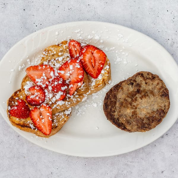 French toast topped with strawberries and powdered sugar, served with two sausage patties on a white plate.