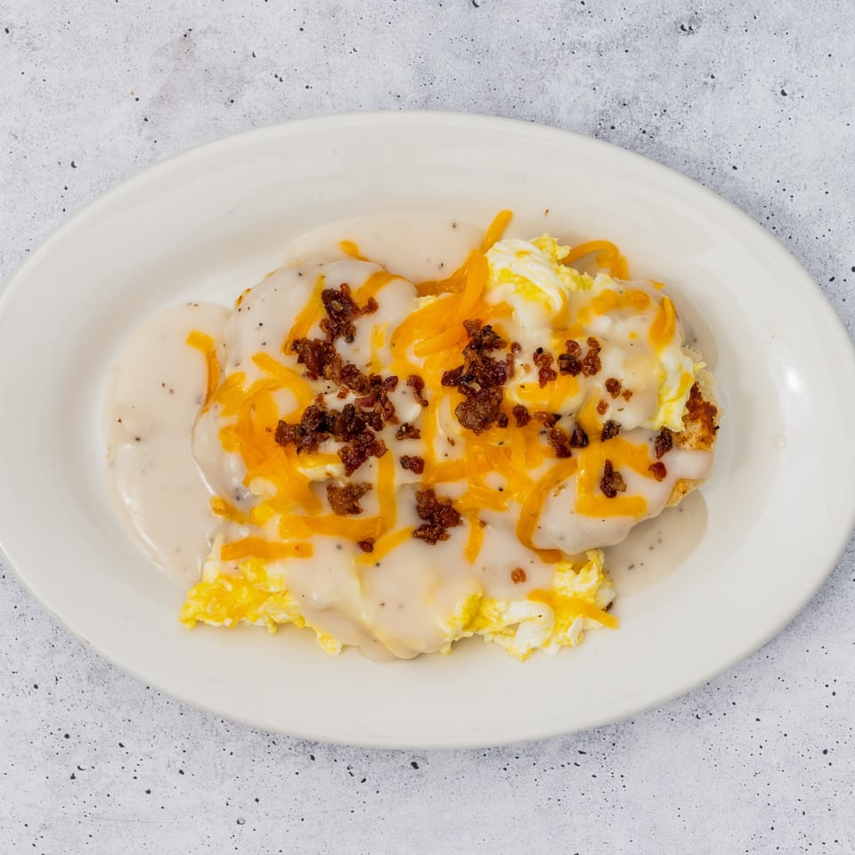 Biscuits and gravy with scrambled eggs, shredded cheddar cheese, and bacon bits on a white plate.