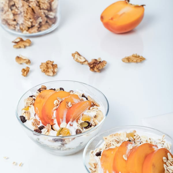 Two bowls of yogurt with sliced persimmon, granola, and nuts, with walnuts and a persimmon half scattered around.