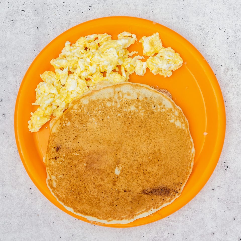A bright orange plate holds fluffy scrambled eggs and a stack of golden pancakes.